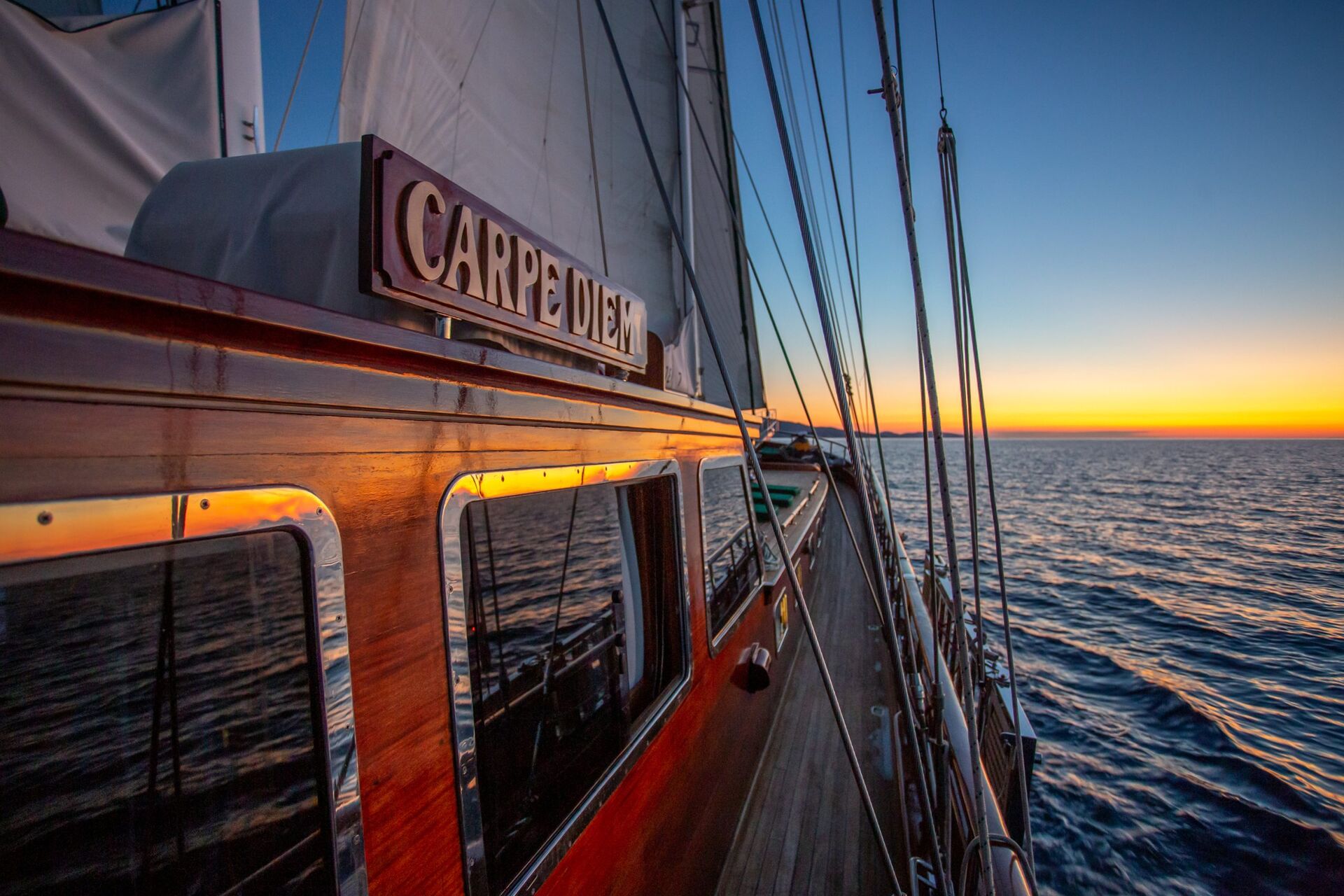 A wooden sailboat named Carpe Diem glides on calm ocean waters at sunset, with warm sunlight reflecting off its windows and sails partially visible.
