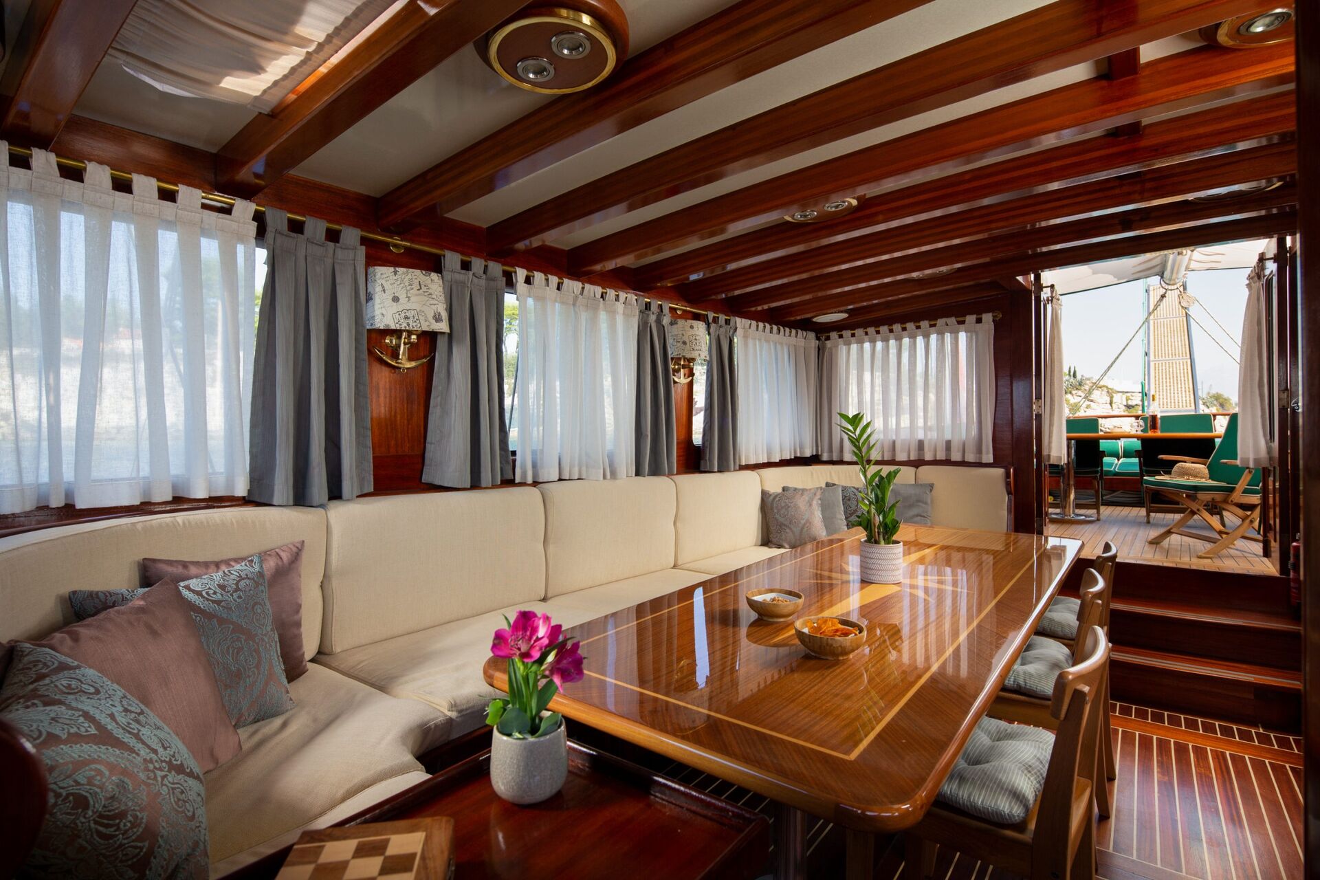 A cozy yacht interior with a wooden ceiling, cream-colored sofa, decorative pillows, a wooden dining table with a potted flower, and large windows with curtains, leading to an outdoor deck with seating.