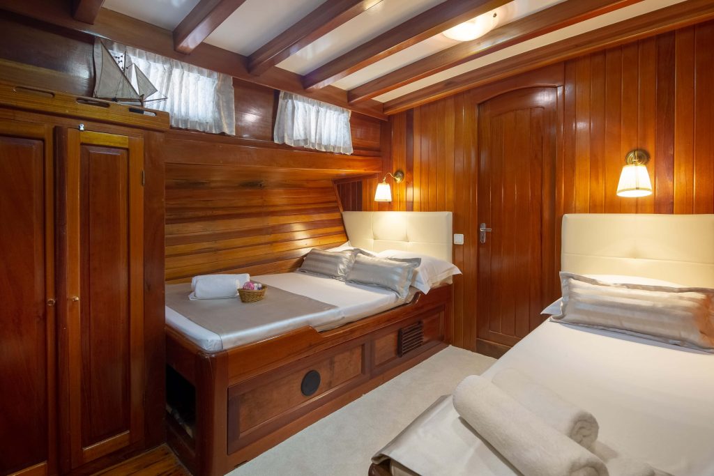 A cozy cabin room with wooden walls and ceiling, two beds with white linens and pillows, wall-mounted reading lights, a wooden door, and a basket with towels on one bed.