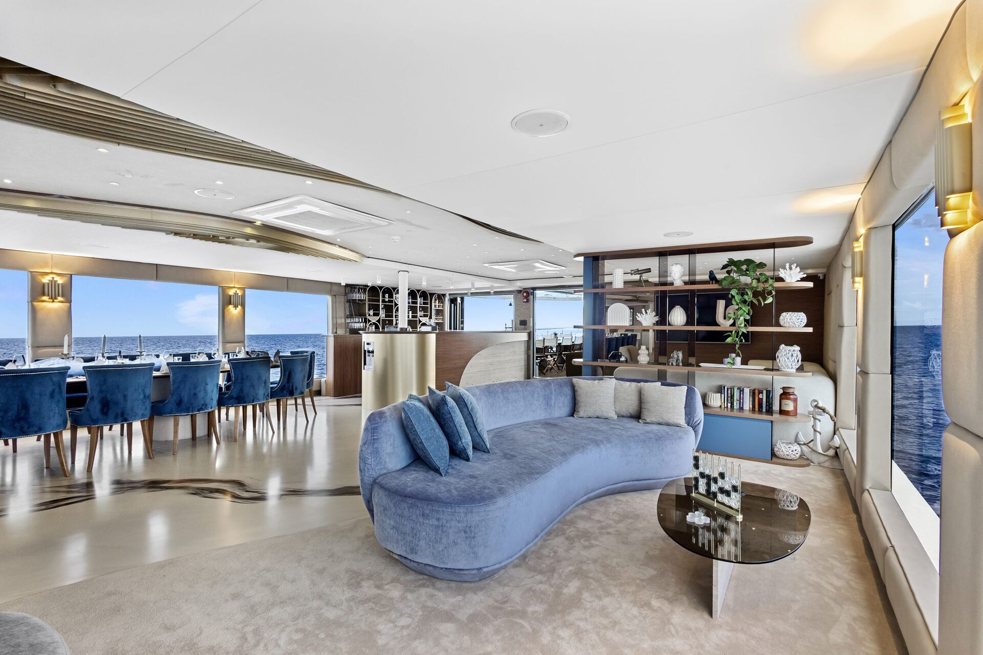 Modern yacht interior with a curved blue sofa, glass coffee table, built-in shelves with decor, large windows showing the ocean, and a dining area in the background with ocean views.
