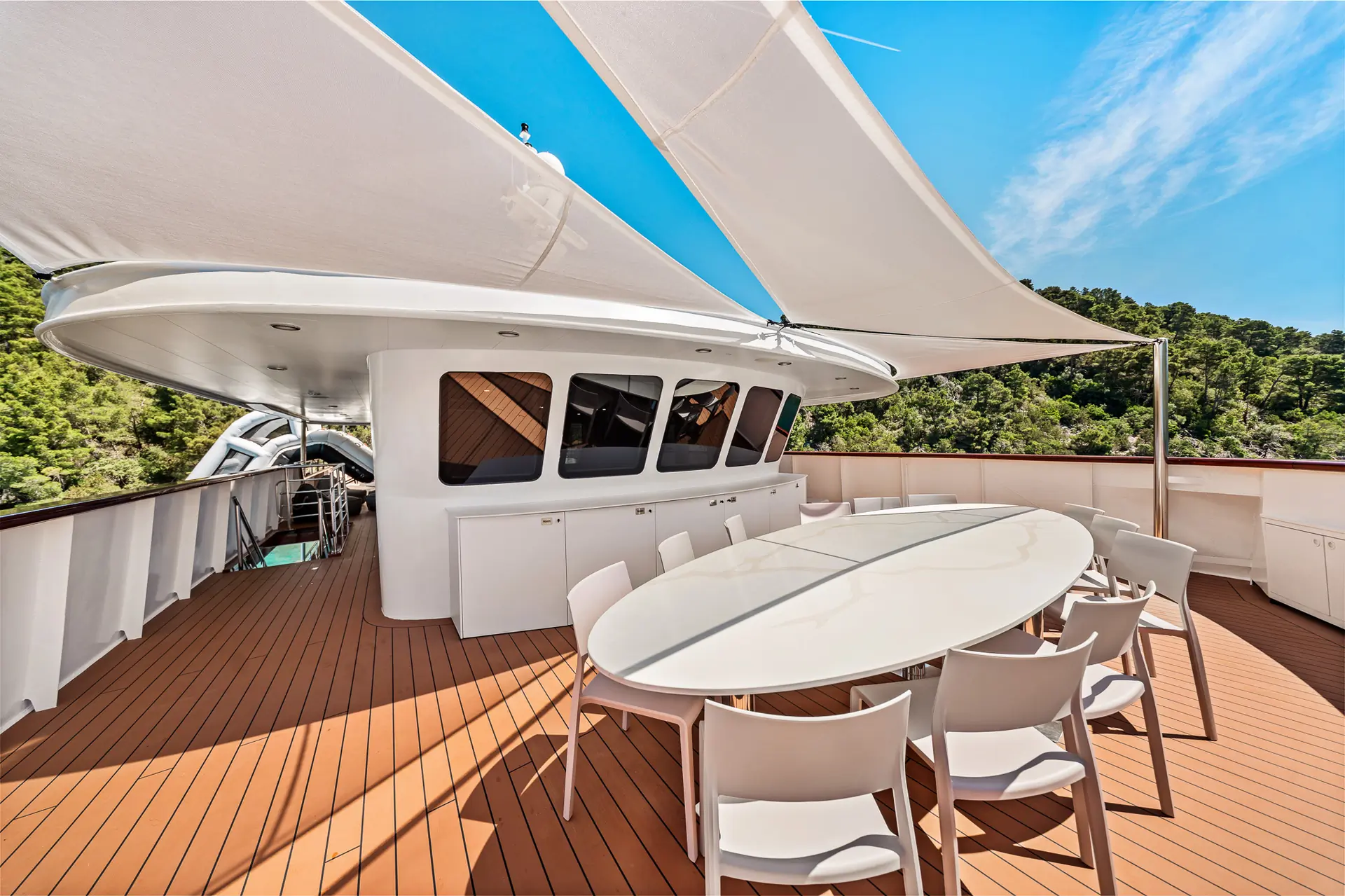 A spacious yacht deck with a large oval dining table and white chairs, shaded by sail-like canopies. The deck features wood-style flooring and is surrounded by greenery and blue sky.