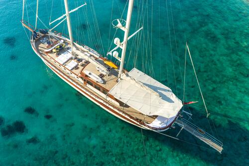 Aerial view of a large sailboat with two masts floating on clear turquoise water. The boat has a wooden deck, white sails, and is anchored near the shore. The sea floor is visible beneath the calm, shallow water.