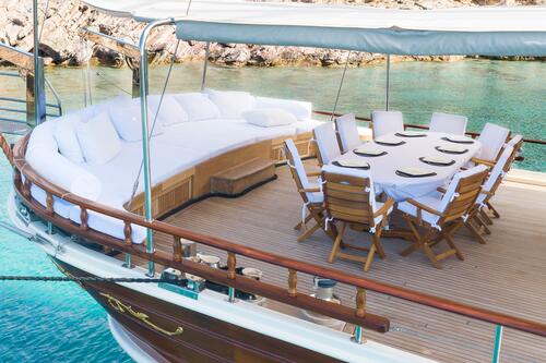 A luxury yacht deck with a curved white cushioned lounge and a dining table set for ten, overlooking clear turquoise water and rocky shoreline in the background.