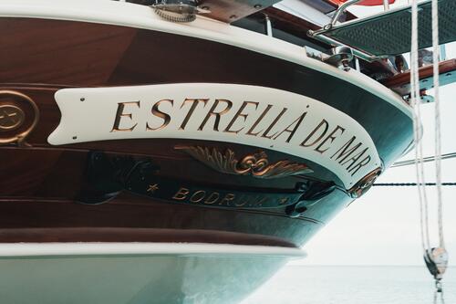 The bow of a wooden boat named ESTRELLA DE MAR with decorative gold lettering and accents is shown. The word BODRUM appears in smaller text below. The sea is visible in the background.