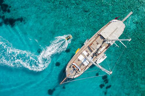 Aerial view of a large sailboat on clear turquoise water, with a small motorboat making a curving wake nearby. Shadows of the boats and underwater features are visible beneath the surface.