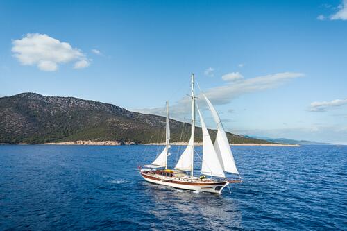 A white sailboat with two masts sails on calm blue water near a forested, hilly coastline under a clear sky with a few clouds.