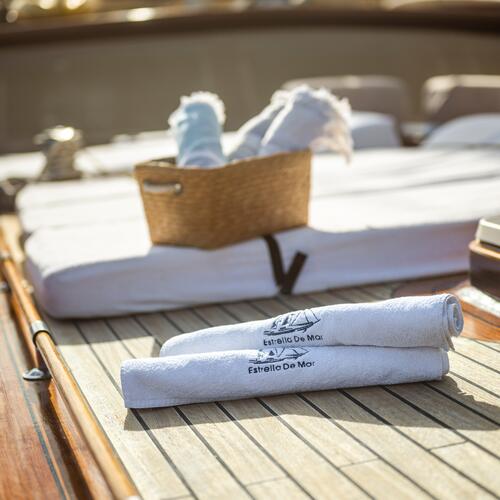Two rolled white towels labeled Estrella De Mar rest on a wooden deck next to loungers. In the background, a wicker basket holds additional rolled towels, all bathed in warm sunlight.