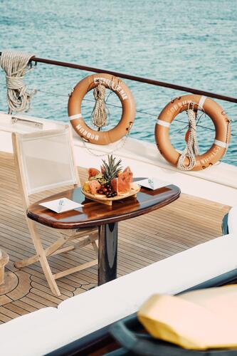 A small wooden table on a boat deck holds a tray of assorted fresh fruit. There is a single chair, and two lifebuoys hang on the railings near calm blue water. Coiled ropes rest nearby.
