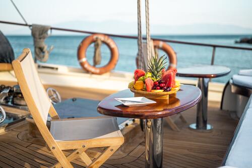 A wooden table with a platter of sliced fruit and a napkin sits next to a deck chair on a yacht, with lifebuoys and ocean water visible in the background.