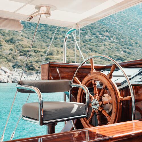 A polished wooden ship’s wheel and a cushioned captain’s chair on a boat deck, with turquoise water and green hills in the background under a canopy.