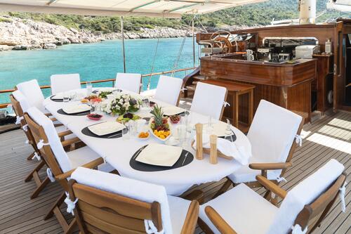 A dining table set for eight with white chairs and a tablecloth on the deck of a yacht, overlooking turquoise water and a rocky, tree-covered shoreline. Food and drink items are arranged on the table.