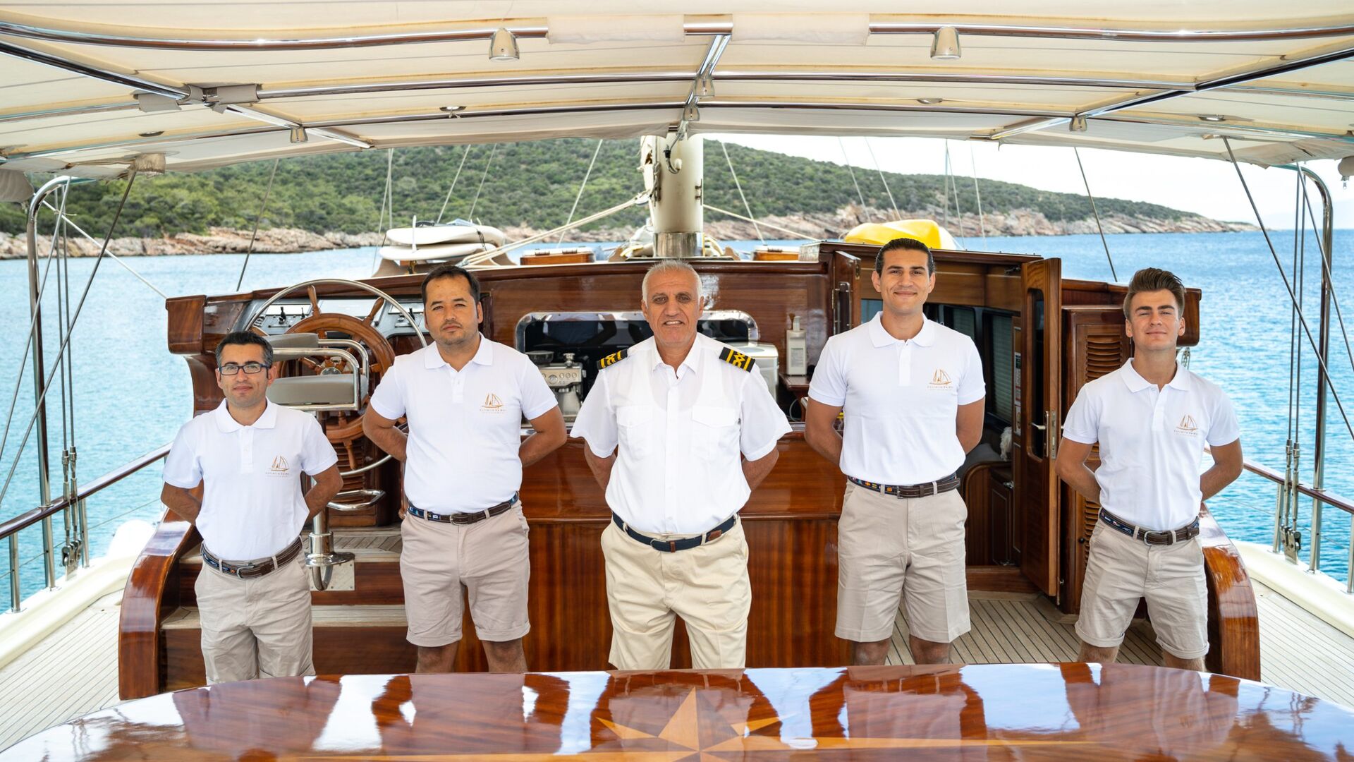 Five crew members in white shirts and beige shorts stand in a row aboard a yacht, with wood-paneled interiors behind them and blue water and green hills visible in the background.