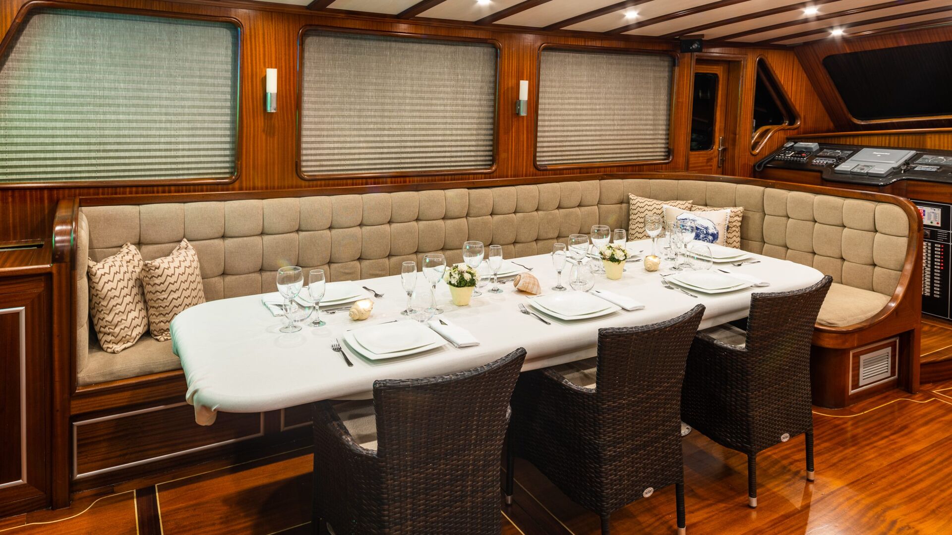 A dining area inside a yacht features a long table set for six, cushioned bench seating along the wall, woven chairs, and wood-paneled walls with closed blinds and nautical-themed decor.
