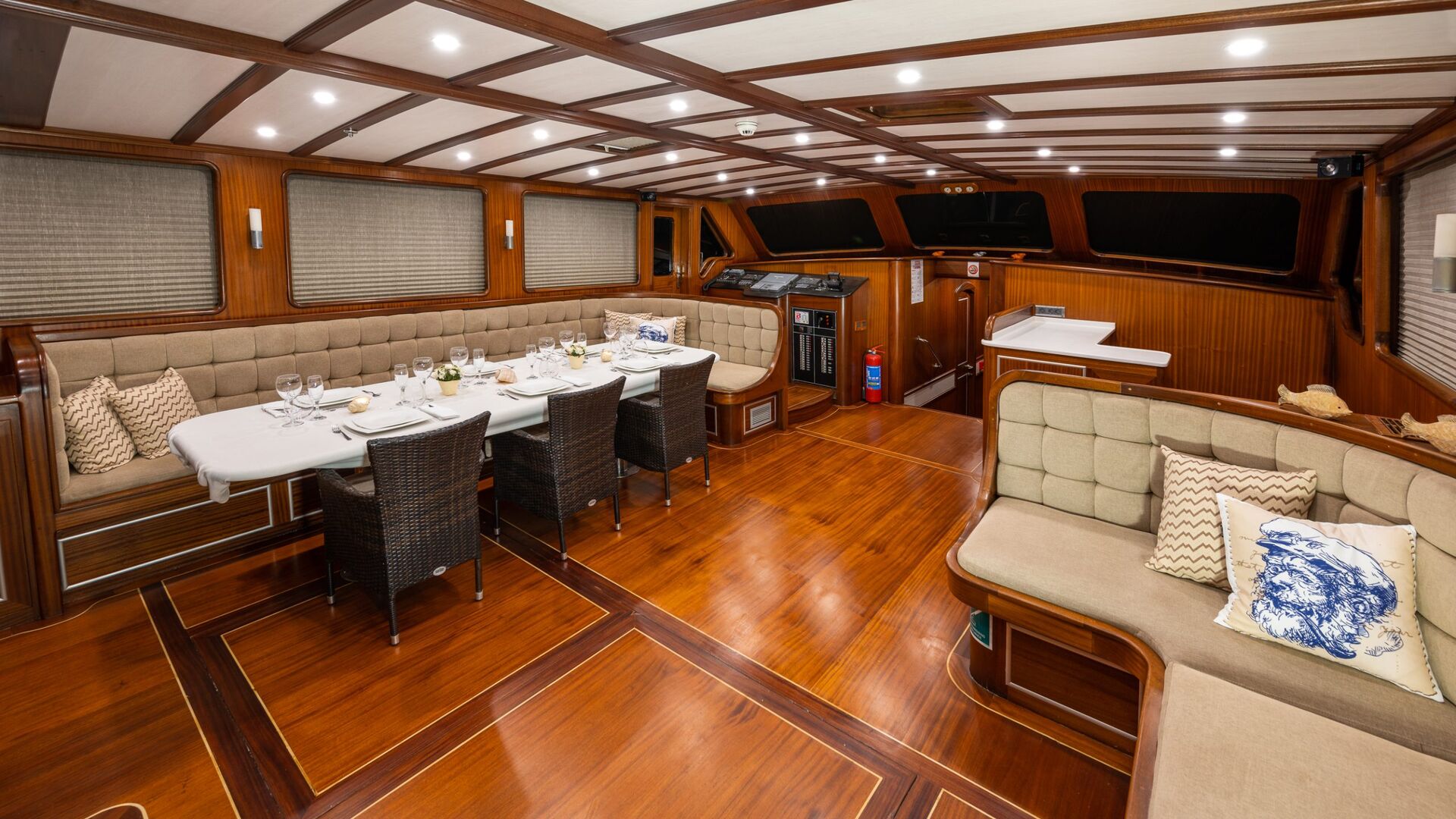 Luxurious yacht interior with polished wood floors, beige cushioned seating, a dining table set for six, large windows with blinds, and ambient ceiling lights, creating an elegant and inviting atmosphere.