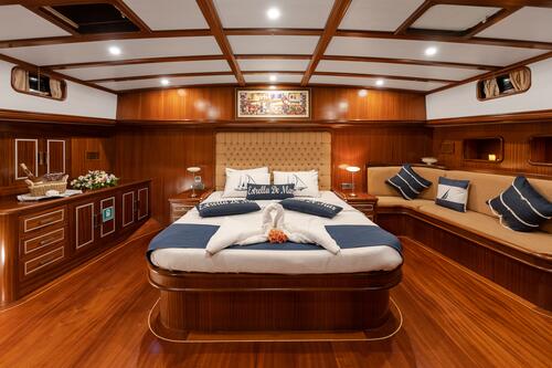 Luxurious yacht bedroom with polished wood walls and floor, a large bed with white and navy bedding, decorative pillows, a built-in sofa, bedside lamps, and nautical-themed decor.