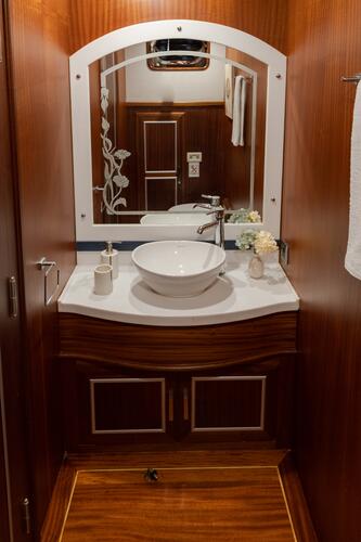 A modern bathroom with wood-paneled walls, a white countertop, a round vessel sink, a silver faucet, a large mirror, and decorative flowers beside the sink. A white towel hangs to the right.