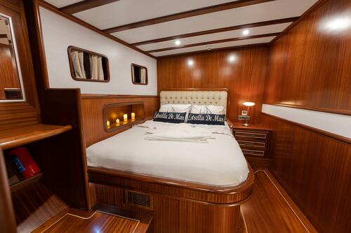 A cozy yacht cabin with polished wood walls and floors, a large bed with white linens and decorative pillows, small windows, soft lighting, and built-in shelves for storage.
