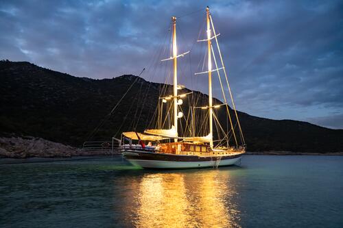 A brightly lit sailboat is anchored in calm, shallow water near a rocky shoreline at dusk, with dark hills in the background and reflections of the boat’s lights shimmering on the water.