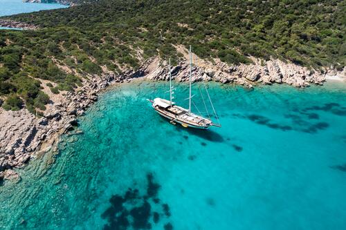 A sailboat is anchored in clear turquoise water near a rocky, forested coastline. The shoreline curves around the bay, and the sea gradually deepens from light blue to darker shades.
