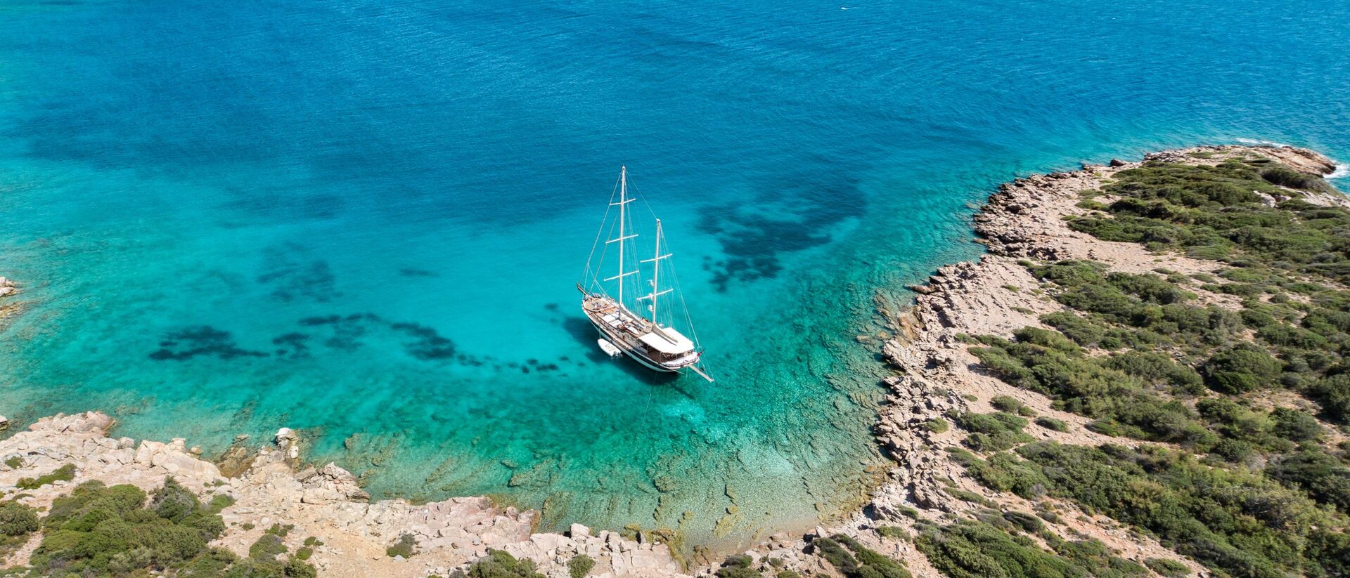 Aerial view of a sailboat anchored in clear turquoise water near a rocky, green-covered coastline with patches of shallow and deep blue sea.