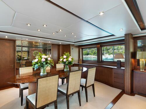 Elegant dining room with a polished wooden table, cream-colored cushioned chairs, and large vases of flowers. Large windows reveal a scenic view of water and greenery. The space is bright, modern, and luxurious.