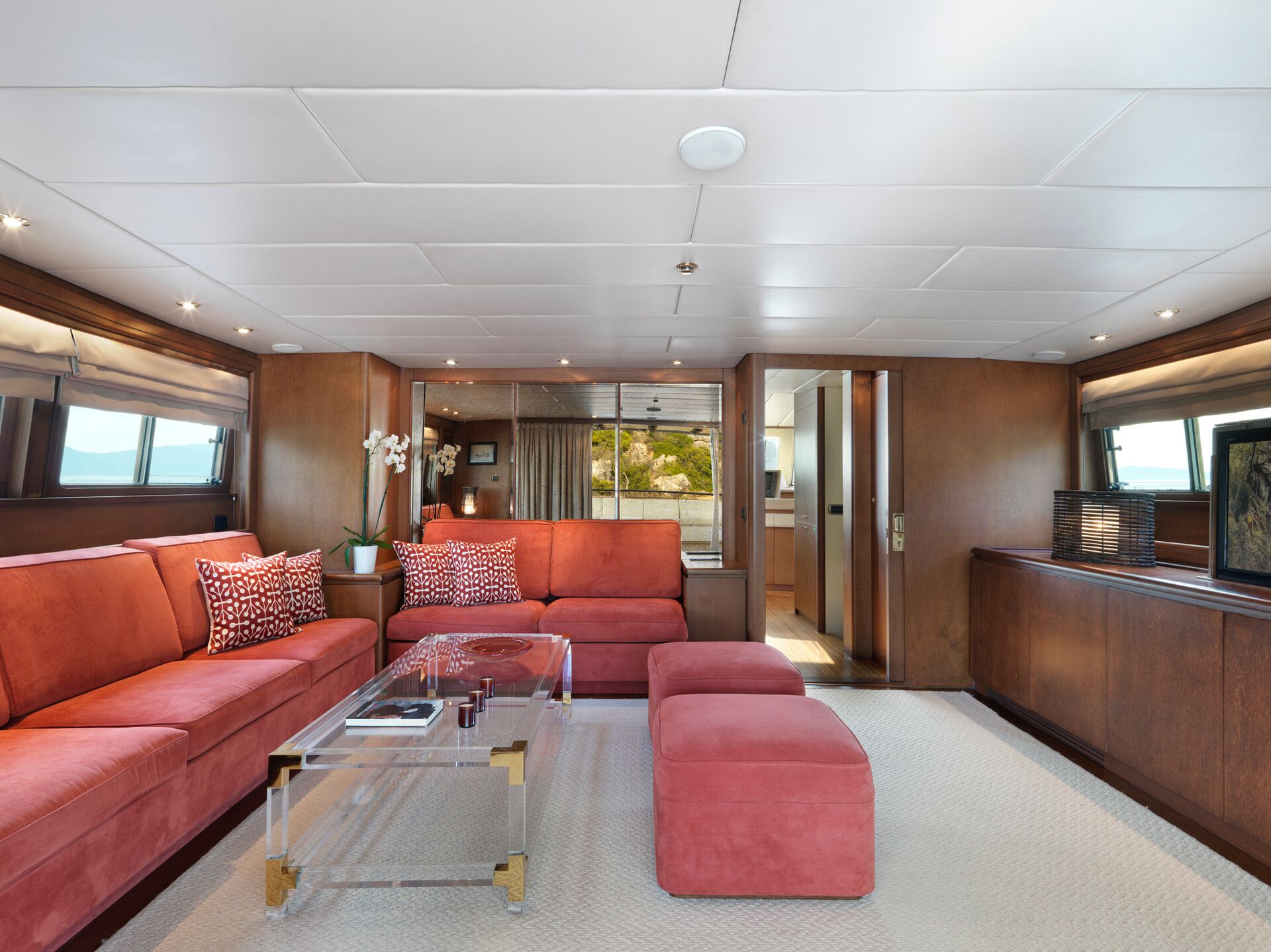 A modern yacht lounge with coral-colored sofas, a clear glass coffee table, and matching ottomans. Large windows provide natural light, and there are orchids and patterned pillows as accents. Wood paneling and neutral carpet complete the space.