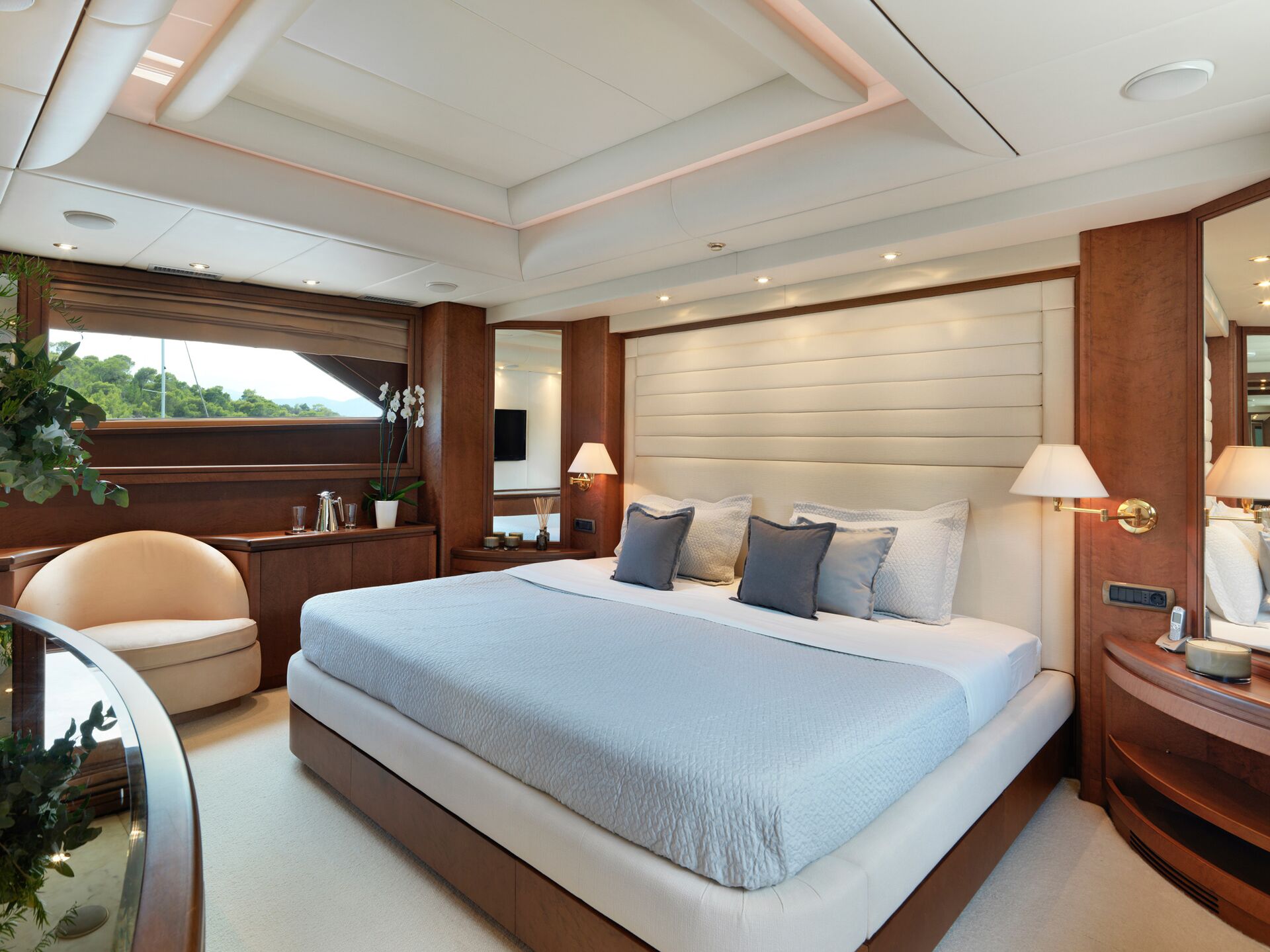 A modern yacht bedroom with a large bed, three blue accent pillows, wood paneling, wall-mounted lamps, a round beige chair, and a window overlooking greenery. The room is brightly lit with a clean, elegant design.