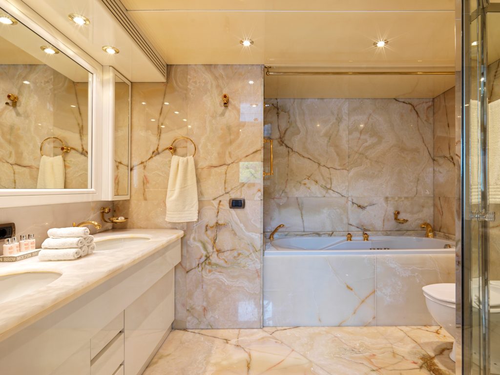 Luxurious bathroom with marble walls and floors, a large bathtub, double sinks with mirrors, gold fixtures, and neatly folded white towels on the counter and towel racks. The space is well-lit with recessed lighting.