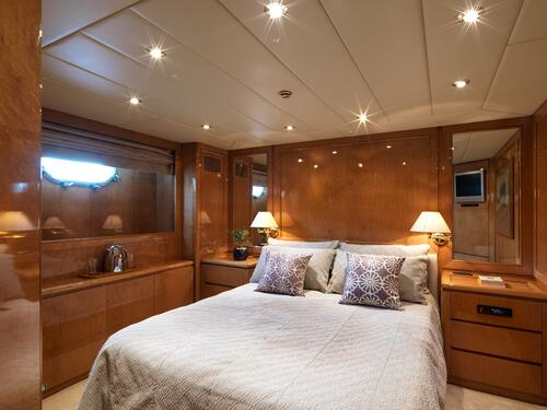A cozy yacht bedroom with wood-paneled walls, a neatly made bed with decorative pillows, bedside tables with lamps, a wall mirror, a small TV, and a window letting in natural light.