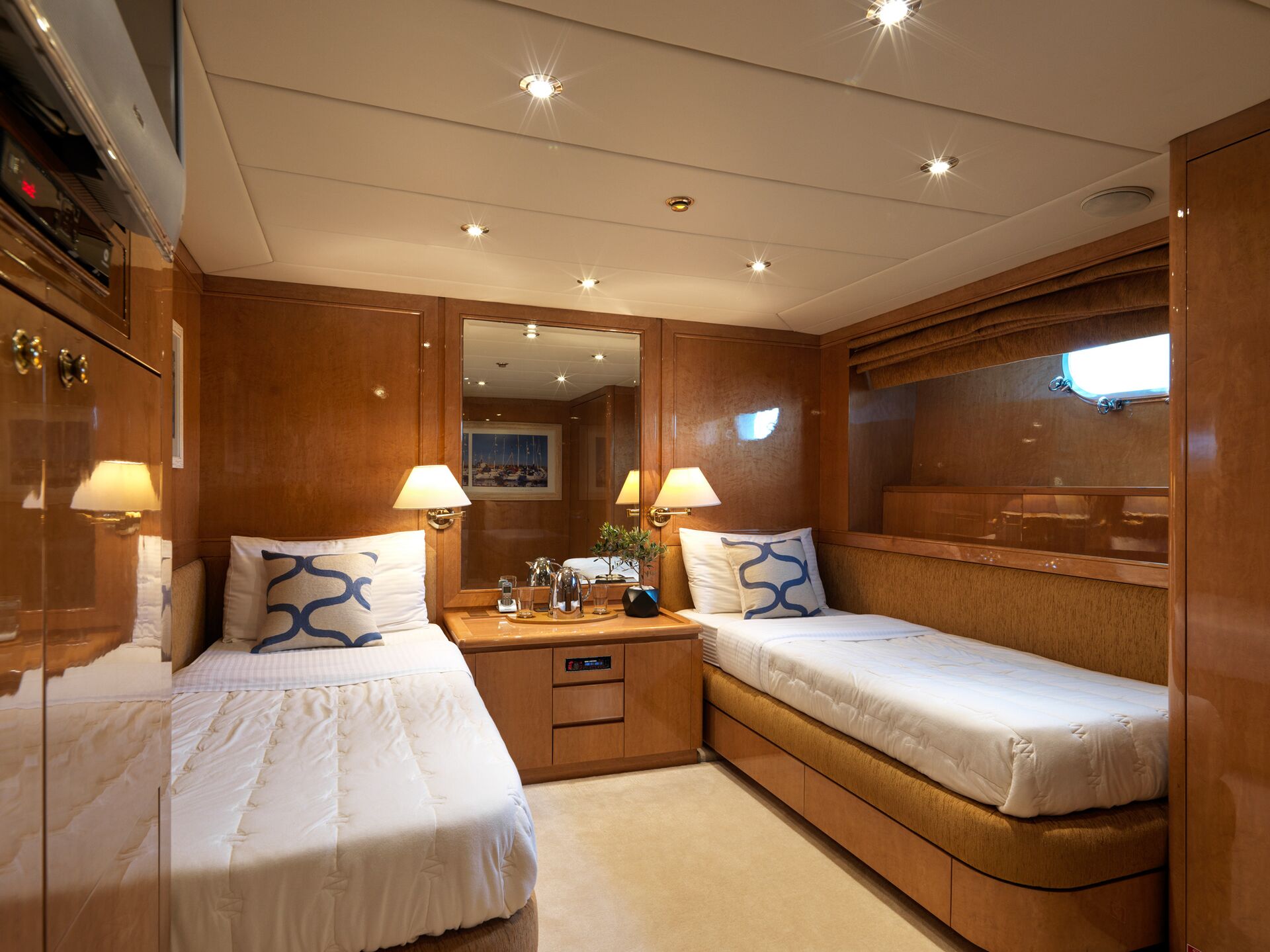 A cozy yacht cabin with wood-paneled walls, two single beds with white linens, a nightstand in between, lamps, a mirror, and a small window with a brown shade.