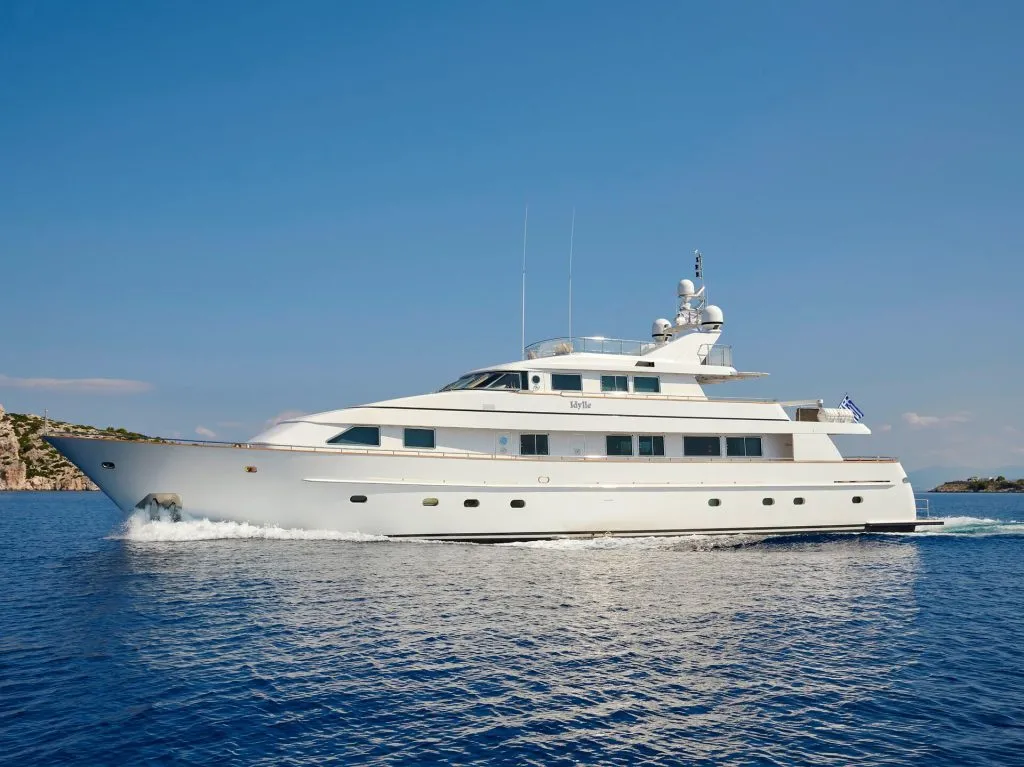 A large white luxury yacht sails across calm blue water under a clear sky, with a small rocky coastline visible in the background.