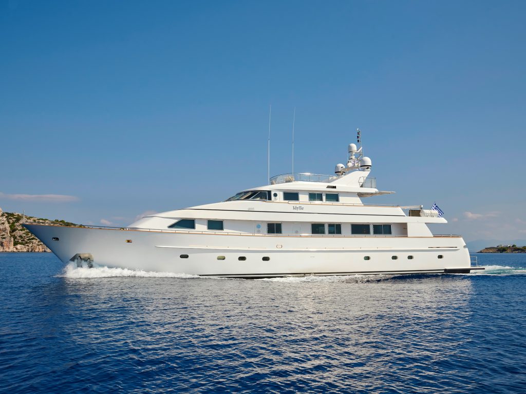 A large white luxury yacht sails across calm blue water under a clear sky, with a small rocky coastline visible in the background.