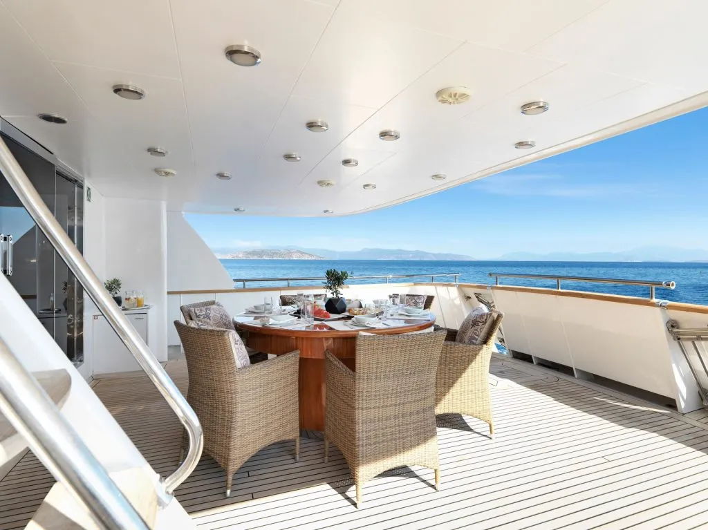 A luxurious yacht deck with a round dining table set for four, wicker chairs, and views of the ocean and distant mountains under a clear blue sky.