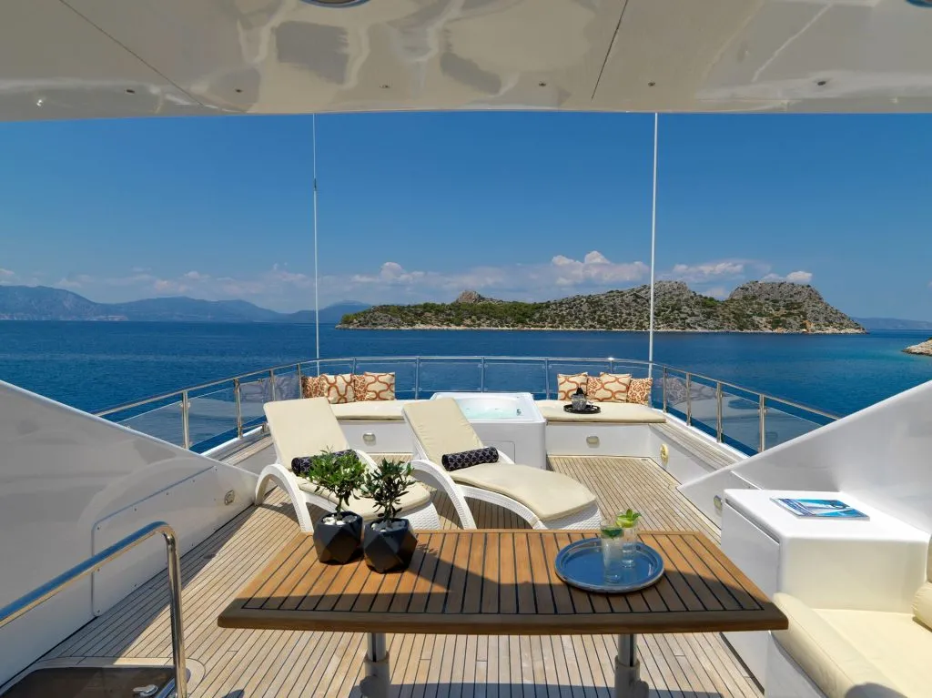 A luxurious yacht deck with sun loungers, a hot tub, and a wooden table overlooks a calm blue sea, with a small green island and distant mountains under a clear sky.