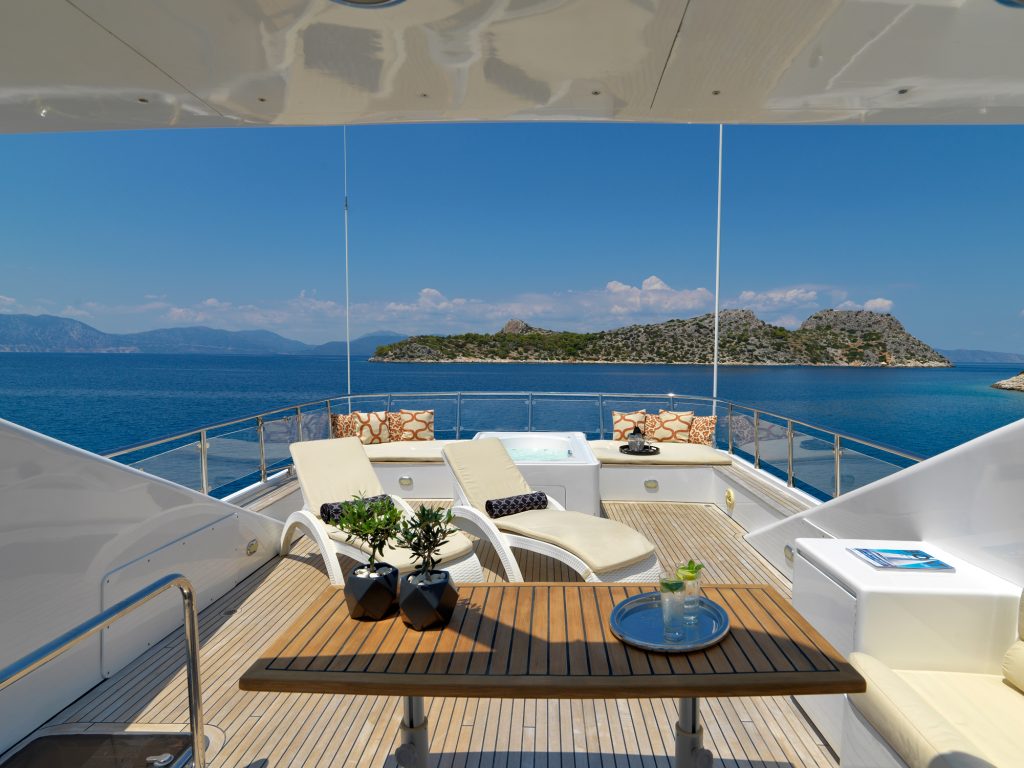A luxurious yacht deck with sun loungers, a hot tub, and a wooden table overlooks a calm blue sea, with a small green island and distant mountains under a clear sky.