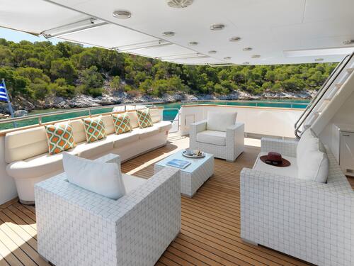 A luxurious yacht deck with white wicker furniture, colorful pillows, a wooden floor, and scenic views of turquoise water and green trees along a rocky shore in the background.