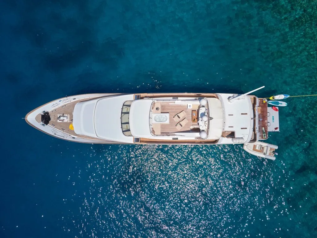 Aerial view of a large white luxury yacht anchored in clear blue water, with a small boat and inflatable water toys floating nearby. The sunlight sparkles on the sea around the yacht.