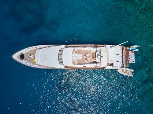 Aerial view of a large white luxury yacht anchored in clear blue water, with a small boat and inflatable water toys floating nearby. The sunlight sparkles on the sea around the yacht.