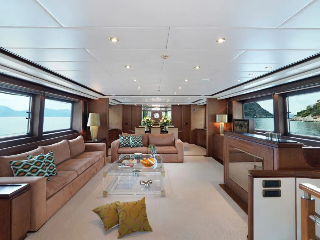 Spacious yacht interior with large windows, tan sofas, colorful pillows, a glass coffee table, and ocean views on both sides. Elegant wooden accents and fresh flowers add a luxurious touch to the room.