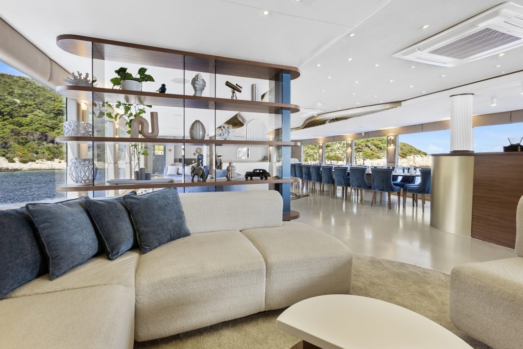 A modern, luxurious yacht interior with cream sofas, blue pillows, a curved open shelf, decorative items, large windows, and an elegant dining area overlooking the water and green hills outside.