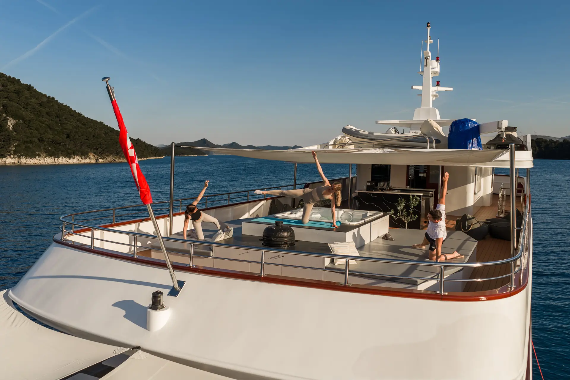 People practicing yoga on the deck of a yacht. The serene scene features individuals in various poses against a backdrop of calm blue water and a lush, green coastline under a clear sky.