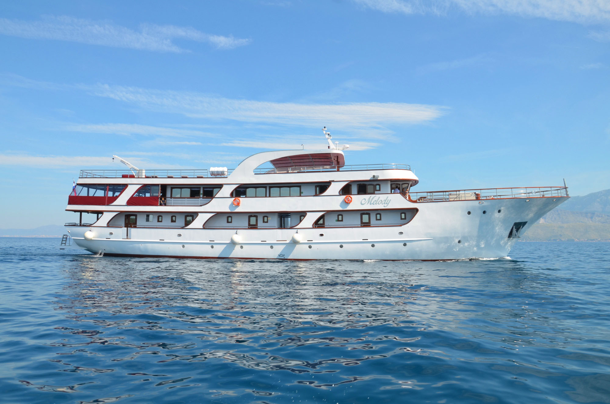 A large white yacht with red accents named Melody floats on calm blue water under a clear sky, with distant mountains visible in the background.