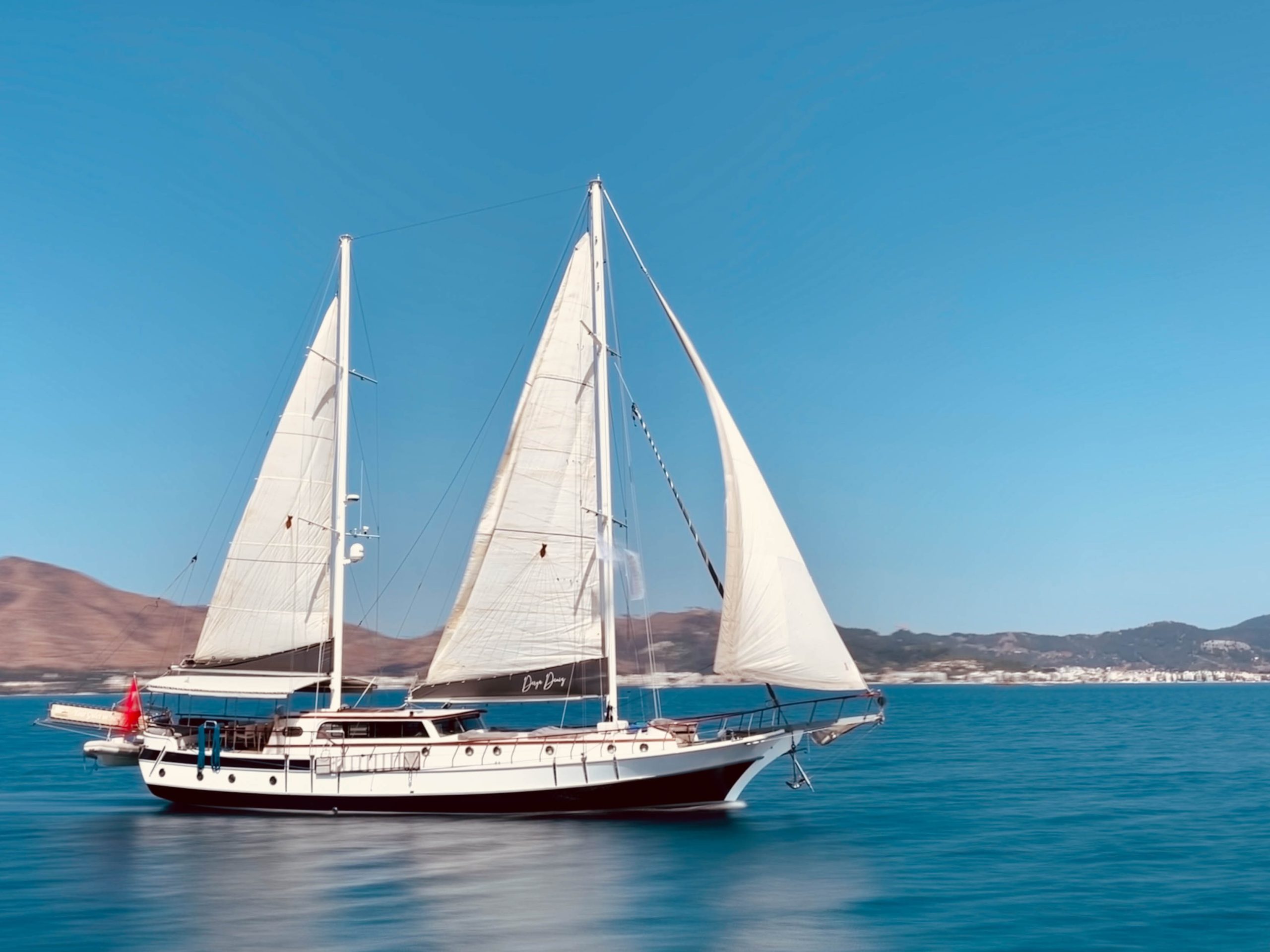A white sailboat with two large sails glides on calm blue water, with hills and a distant shoreline visible under a clear, bright sky.