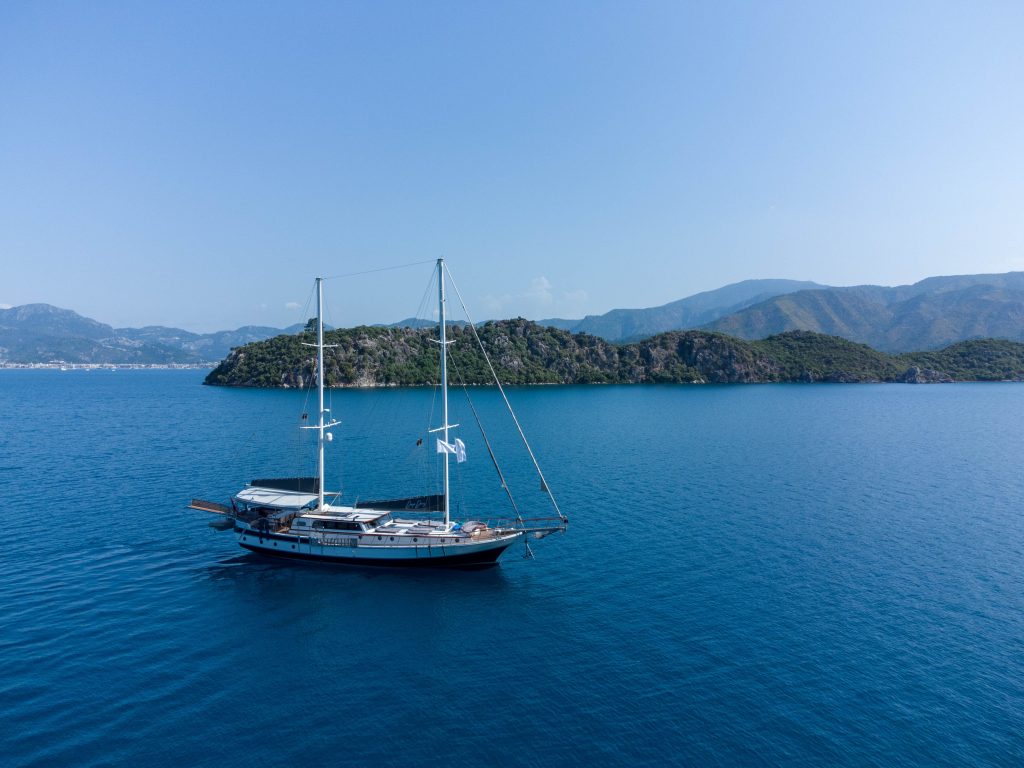 A sailboat with two masts floats on calm blue water near a green, hilly island under a clear sky, with distant mountains visible in the background.