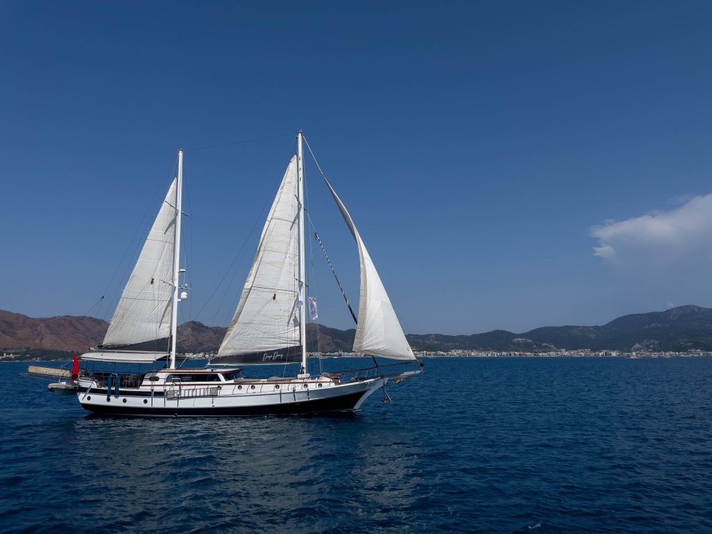 A white sailboat with two sails glides on calm blue water, with a coastal town and mountains visible under a clear blue sky in the background.