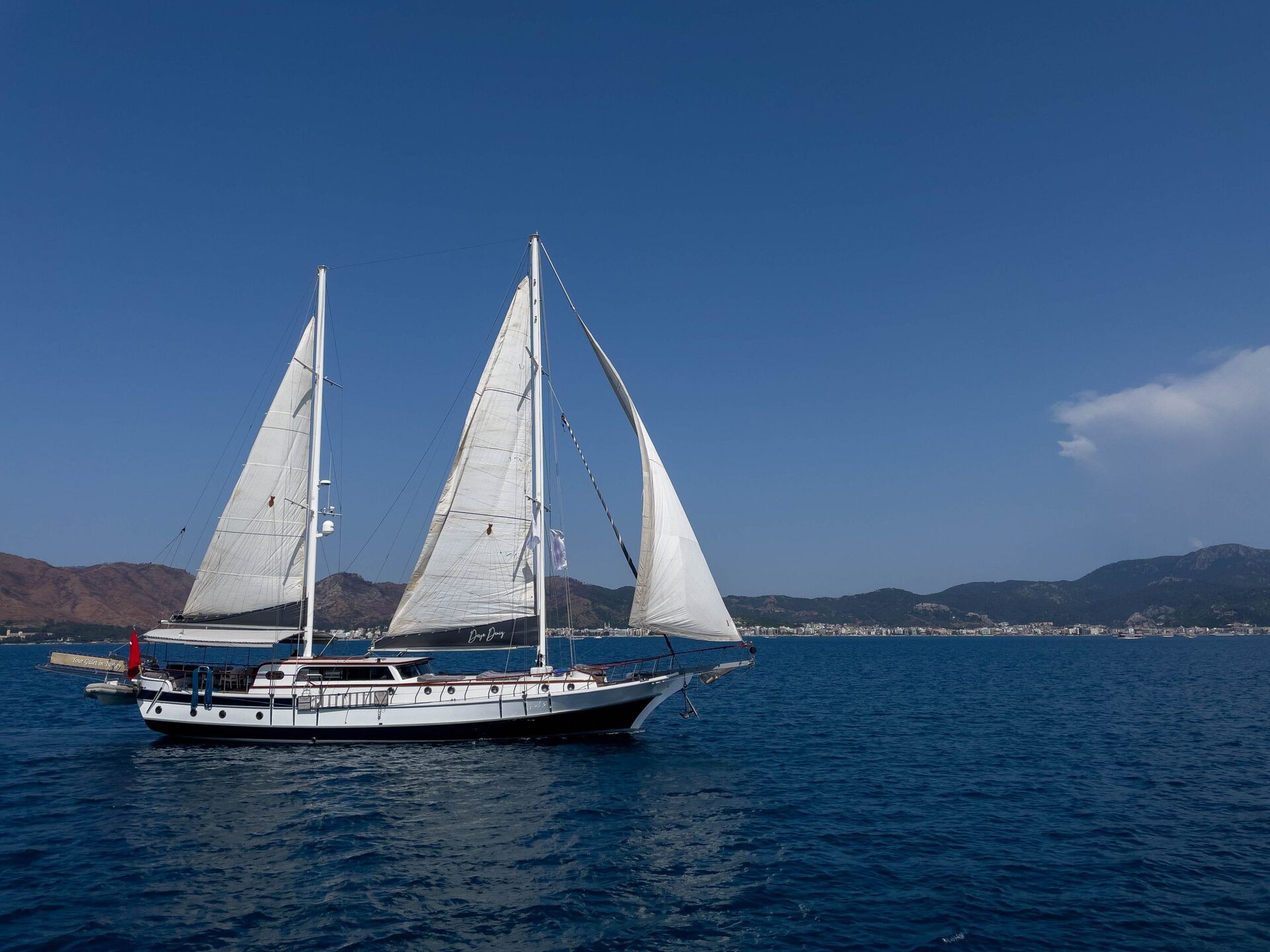A white sailboat with two sails glides on calm blue water, with a coastal town and mountains visible under a clear blue sky in the background.