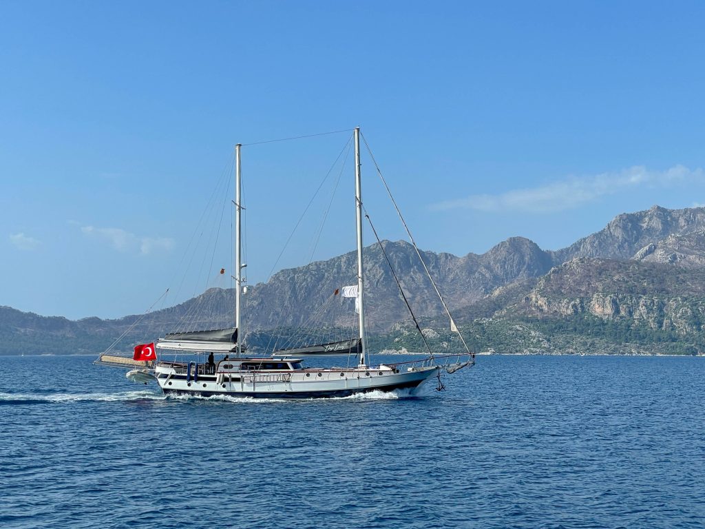 A white sailboat with a Turkish flag sails on calm blue water, with rocky mountains and a clear blue sky in the background.