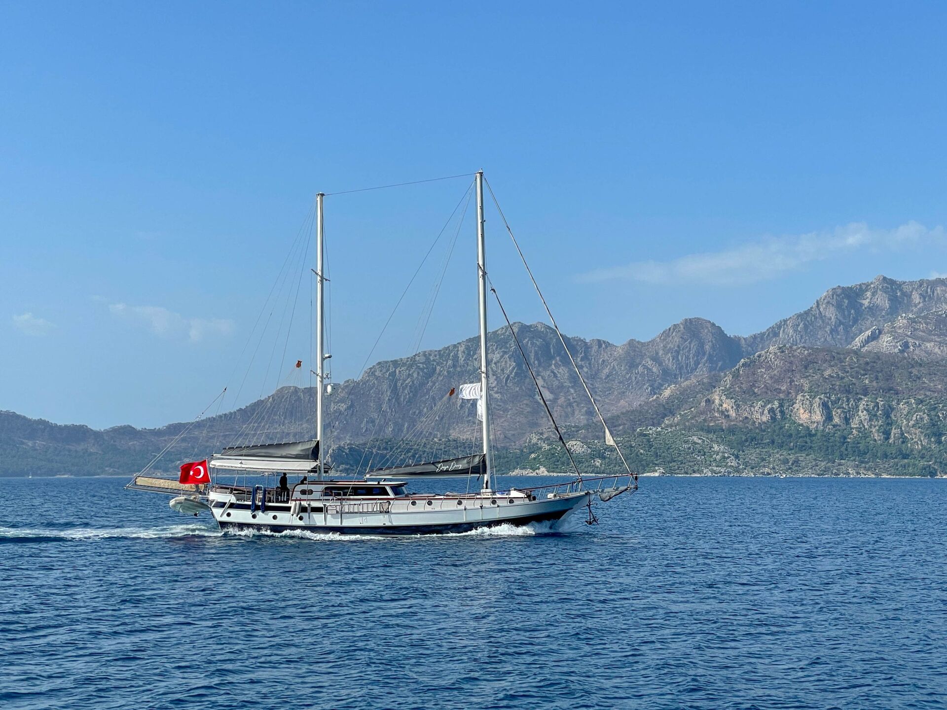 A white sailboat with a Turkish flag sails on calm blue water, with rocky mountains and a clear blue sky in the background.