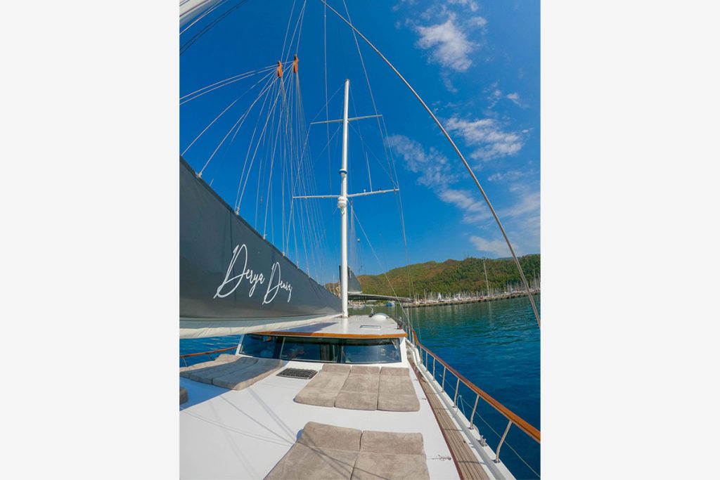 View from the deck of a sailboat with lounge cushions, a large sail bearing the words “Deep Jump,” calm blue water, and green hills under a bright partly cloudy sky.
