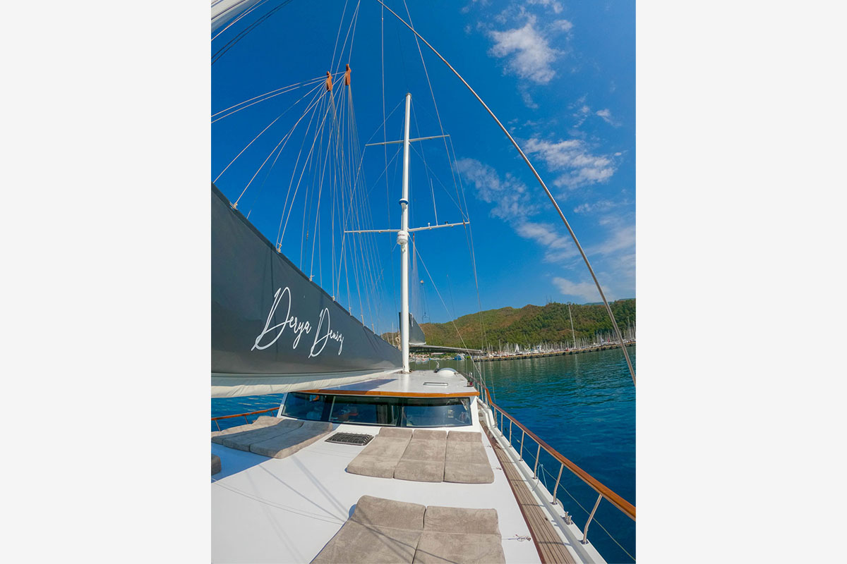 View from the deck of a sailboat with lounge cushions, a large sail bearing the words “Deep Jump,” calm blue water, and green hills under a bright partly cloudy sky.
